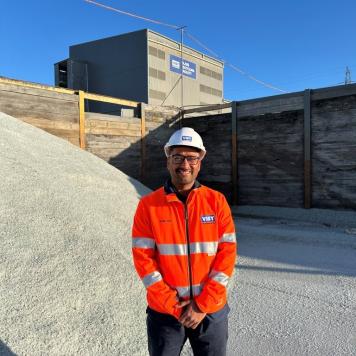 Man smiling at camera in orange high vis work wear with white hard hat. Standing in front of clear recycled glass and a large factory.