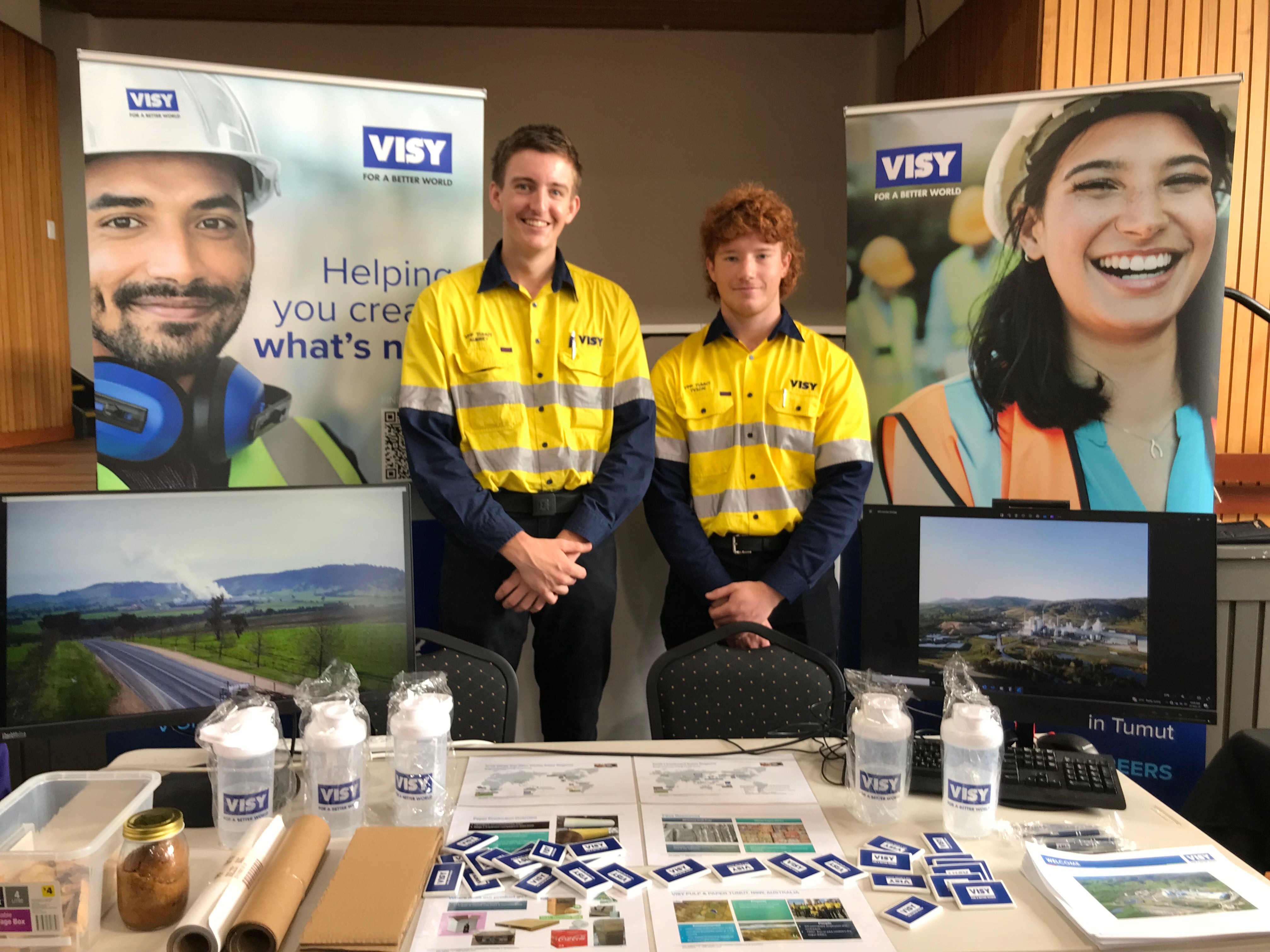 Two young men in high visibility workwear are smiling at the camera, behind a table with Visy branded merchandise 