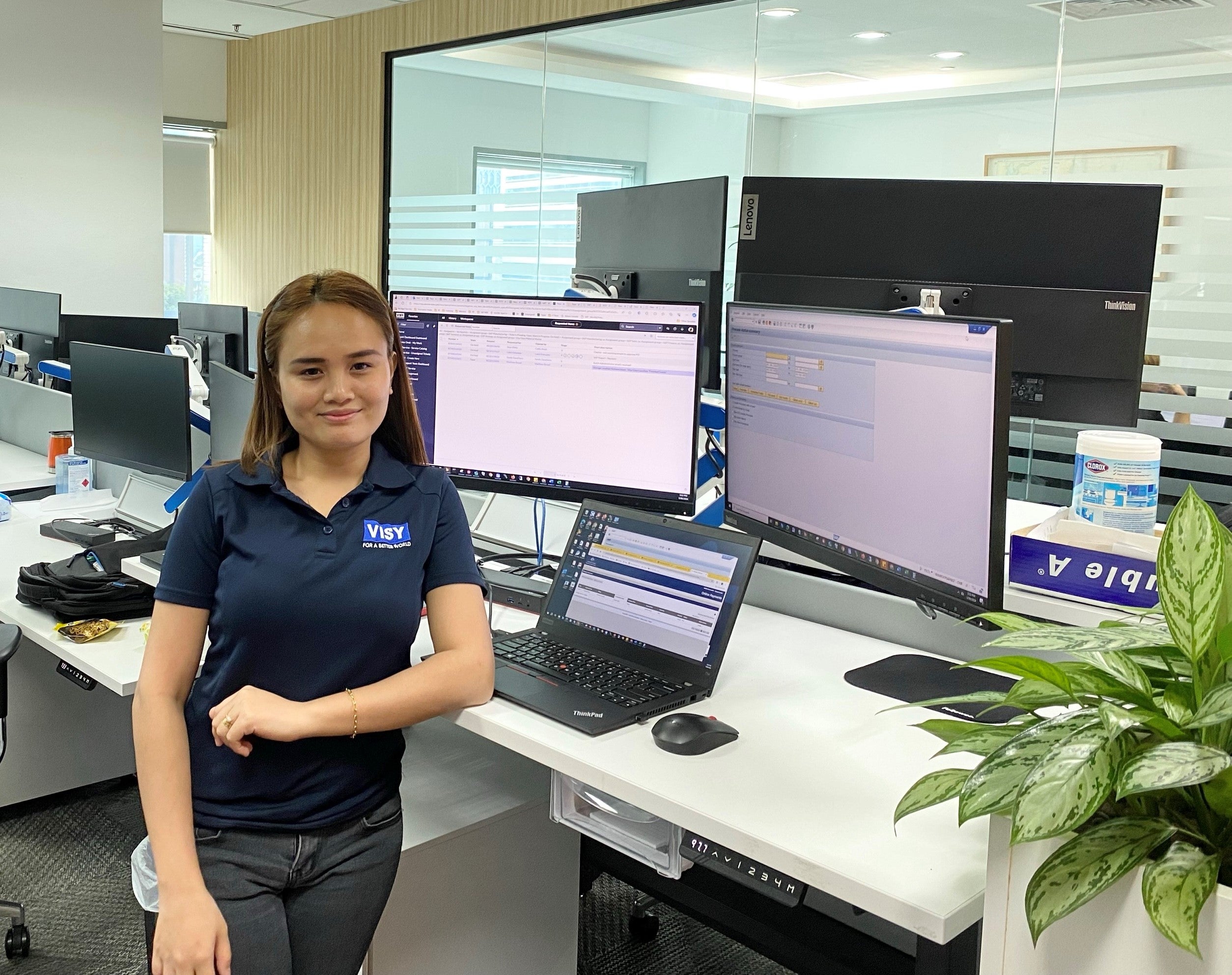 A woman smiles at the camera, wearing a blue Visy-branded polo shirt. She is in an office at a desk with 3 computer screens