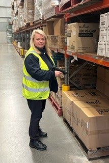 A woman wearing a high vis safety best, holding an inventory scanner in a warehouse full of product looks at the camera