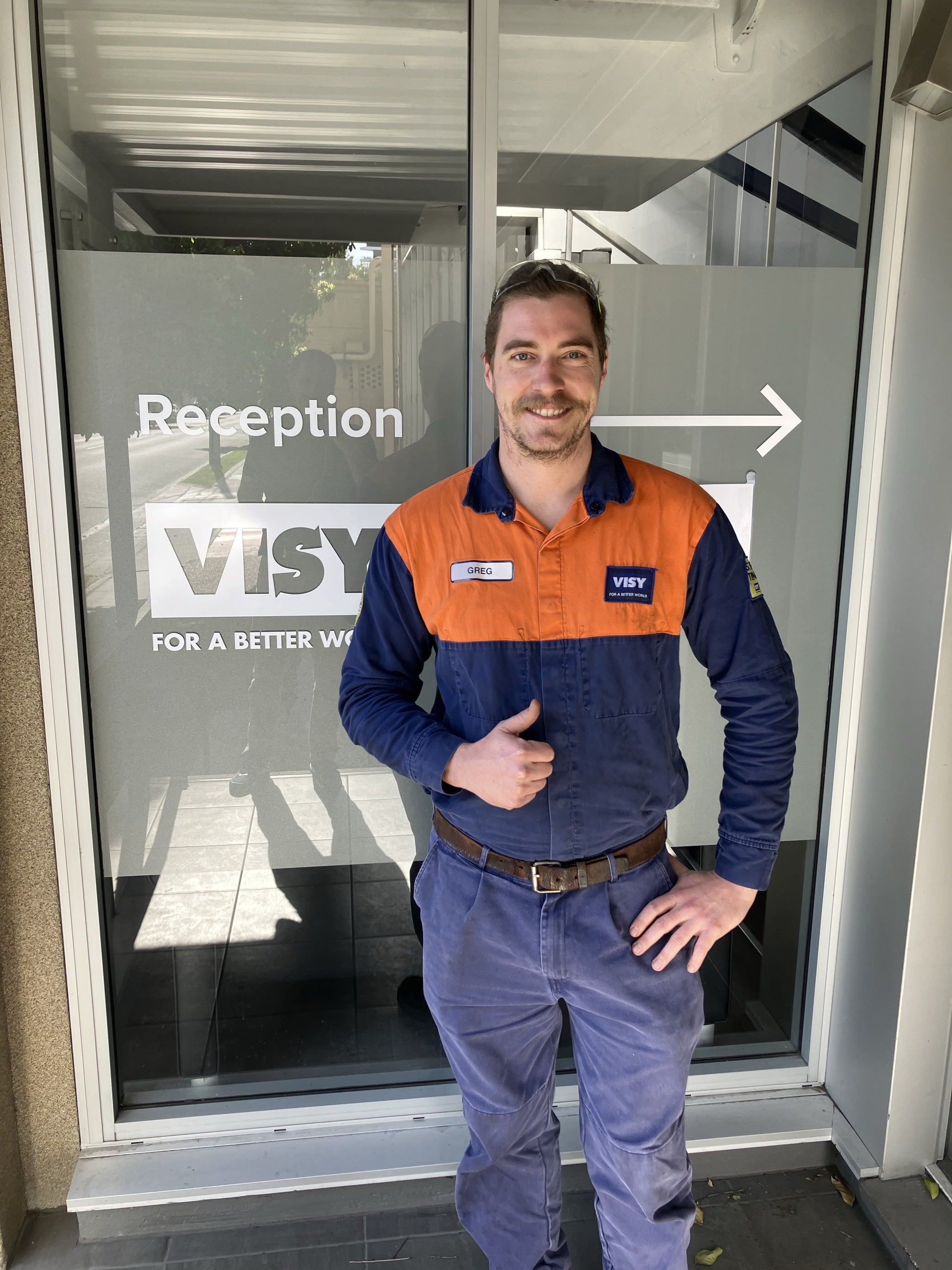 A man smiles at the camera, in blue and orange safety work wear standing in front of a building entrance giving a thumbs up