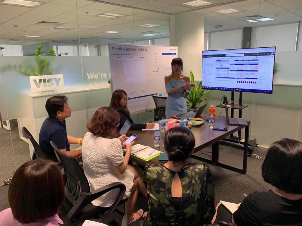 A woman in a blue dress is speaking to 5 people in an office about a presentation on screen beside her. 