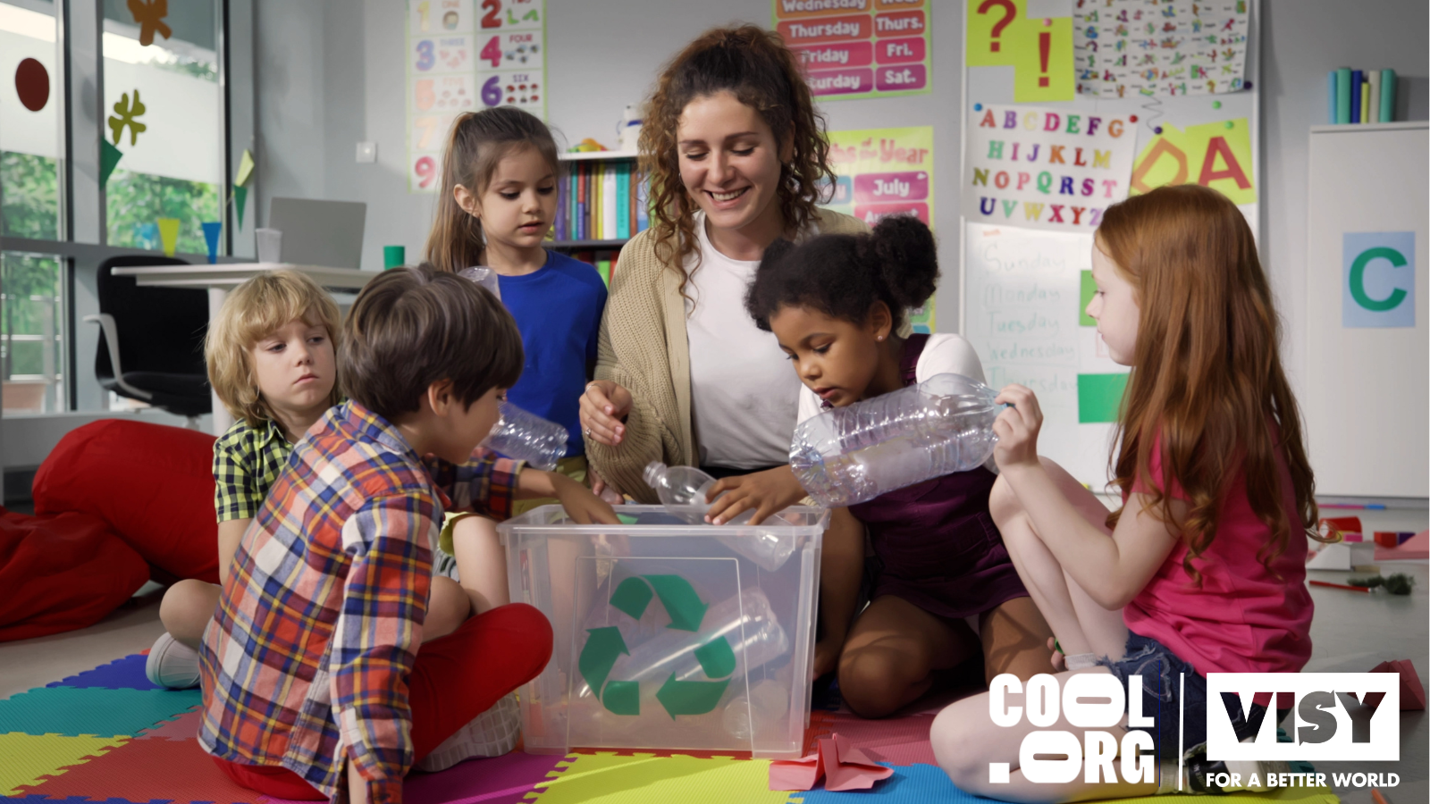 6 children putting plastic bottles into a clear recycling bin with the help of their teacher