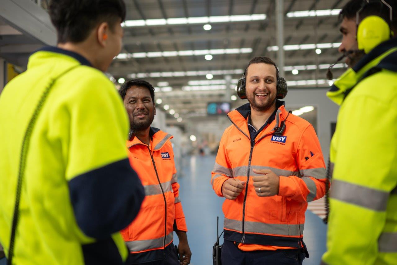 Two men in orange high vis tops are smiling towards the camera talking to two people with their backs to camera