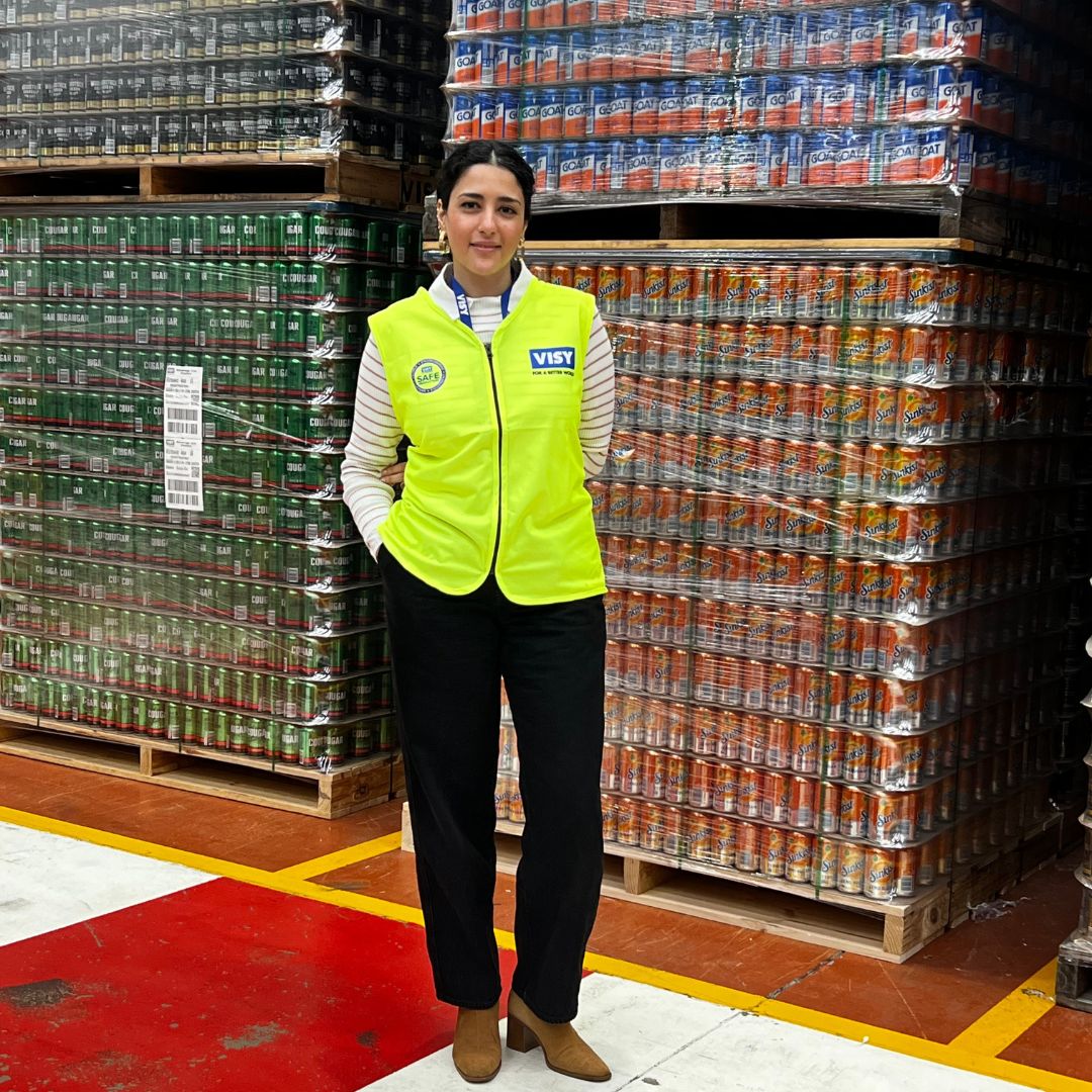 A woman with dark hair in a yellow high visibility safety vest smiles at camera, she's standing in front of aluminium beverage cans