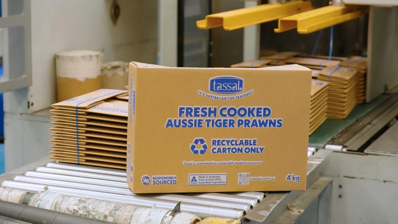 A brown cardboard box with blue writing on it sits on a conveyor belt with stacks of flattened boxes behind it