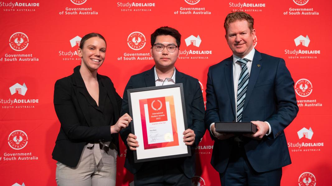 Three people stand in front of a red marketing banner holding a framed certificate