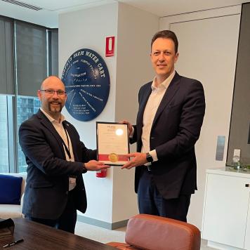 Two men in suits, in an office, smiling and facing the camera, they are holding a framed certificate between them