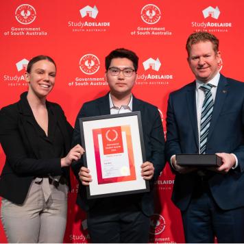 Three people stand in front of a red marketing banner holding a framed certificate