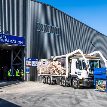 A front loading truck with a bin of cardboard for recycling in front of a shed