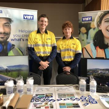 Two young men in high visibility workwear are smiling at the camera, behind a table with Visy branded merchandise 