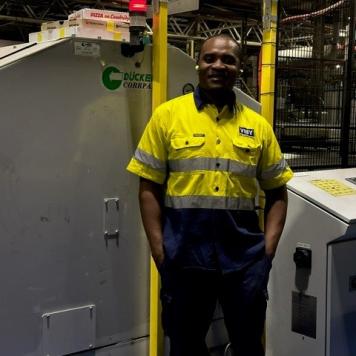 A male employee in a high vis safety shirt smiles at the camera, he is standing in front of machinery