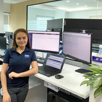 A woman smiles at the camera, wearing a blue Visy-branded polo shirt. She is in an office at a desk with 3 computer screens