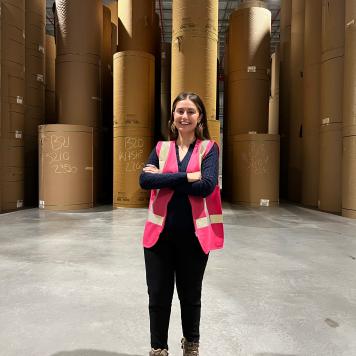 A woman smiles at the camera, she's wearing a pink hi vis safety vest and dark clothes. She's in a warehouse with brown paper reels