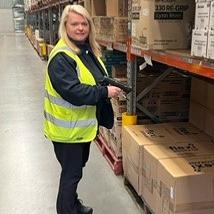 A woman wearing a high vis safety best, holding an inventory scanner in a warehouse full of product looks at the camera