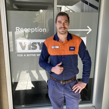 A man smiles at the camera, in blue and orange safety work wear standing in front of a building entrance giving a thumbs up