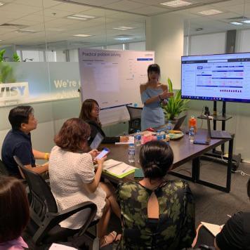 A woman in a blue dress is speaking to 5 people in an office about a presentation on screen beside her. 