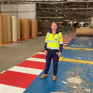 A woman in high vis safety gear smiles at the camera. She is in a warehouse with brown paper reels behind her. 