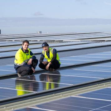 Two men in high visibility safety vests are squatting on a roof, surrounded by solar panels