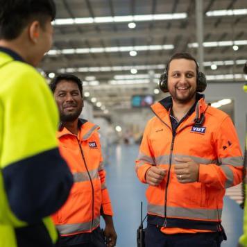 Two men in orange high vis tops are smiling towards the camera talking to two people with their backs to camera