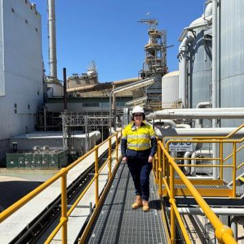 A woman smiles at camera. She is wearing high visibility, safety work wear and a white hard hat. She is surrounding by industrial looking pipes and building. The sky is blue. 