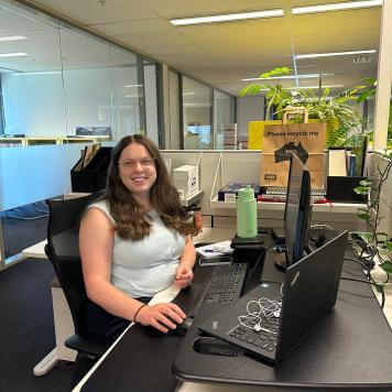 A young woman smiles at the camera, sitting in an office at a computer, she is wearing a light top