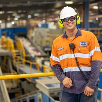 A man in safety gear smiles at the camera, he is in an industrial looking building