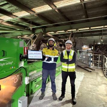 Two men in high visibility safety wear are smiling at the camera with arms folded, they are in a warehouse with equipment behind them