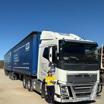 A woman in yellow high visibility work wear smiles with a thumbs up to the camera, she is standing in front of a large blue truck
