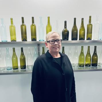 Woman in glasses and dark top smiles at the camera with a wall of glass bottles behind her.
